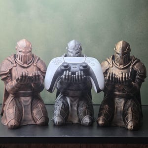 Peut inclure: Trois figurines de chevaliers agenouillés, les mains tendues vers l'avant. Le chevalier central tient une manette de jeu vidéo blanche et noire. Les personnages présentent des finitions métalliques cuivrées, argentées et bronzées avec une armure détaillée.