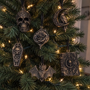 May include: A set of six hanging ornaments in a dark bronze finish, featuring a skull, a crescent moon with a raven, a rose, a coffin with a skull, a bat, and a book with a skull emblem. Each piece is suspended by a black ribbon on a pine tree.