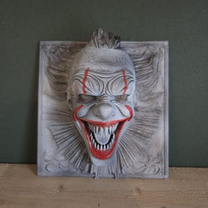 May include: A gray, 3D-printed clown face with red accents. The clown has a wide, menacing grin with sharp teeth and red lines painted on the face. The clown's hair is black and spiky, and the face is set against a gray, textured background.