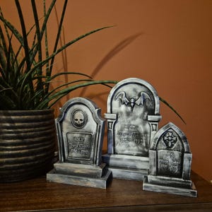 Op de afbeelding: Drie grijze en zwarte Halloween grafstenen met de tekst "RIP Myra Mains 1736-1790". Eén grafsteen heeft een schedel, een andere een vleermuis en de derde een kruis. Een potplant staat op de achtergrond.