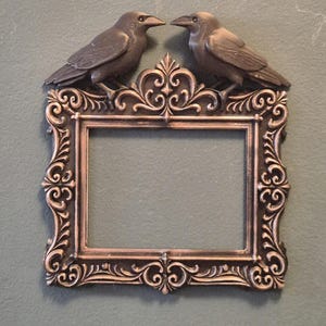 Op de afbeelding: Een sierlijke bronzen fotolijst met twee gebeeldhouwde kraaien erboven. Het frame heeft ingewikkelde krullen en een rechthoekige opening. De kraaien kijken elkaar aan en geven het decor een gotische of vintage uitstraling.