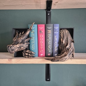May include: Bronze dragon bookends holding a row of books with titles including "Eragon" and "Brisingr". The bookends have a dragon head and wing design. The books are in various colours. The shelf is made of light-coloured wood.