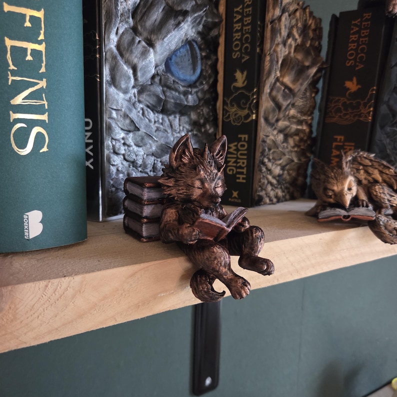 May include: A bronze-colored fox figurine reading a book, perched on a wooden shelf. The fox is sitting on a stack of books. Other decorative bookends are visible, including an owl reading a book. Books with gold lettering are in the background.