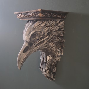 May include: A grey wall-mounted shelf with a sculpted raven skull design. The shelf has a weathered appearance, and the top has a decorative border. The raven's head is detailed with feathers and a long beak, adding a gothic touch.