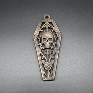 May include: A bronze-toned metal pendant shaped like a coffin, featuring an intricate openwork design. The center displays a detailed human skull positioned above a cross and a blooming rose, all framed by ornate scrollwork and a small hanging loop.