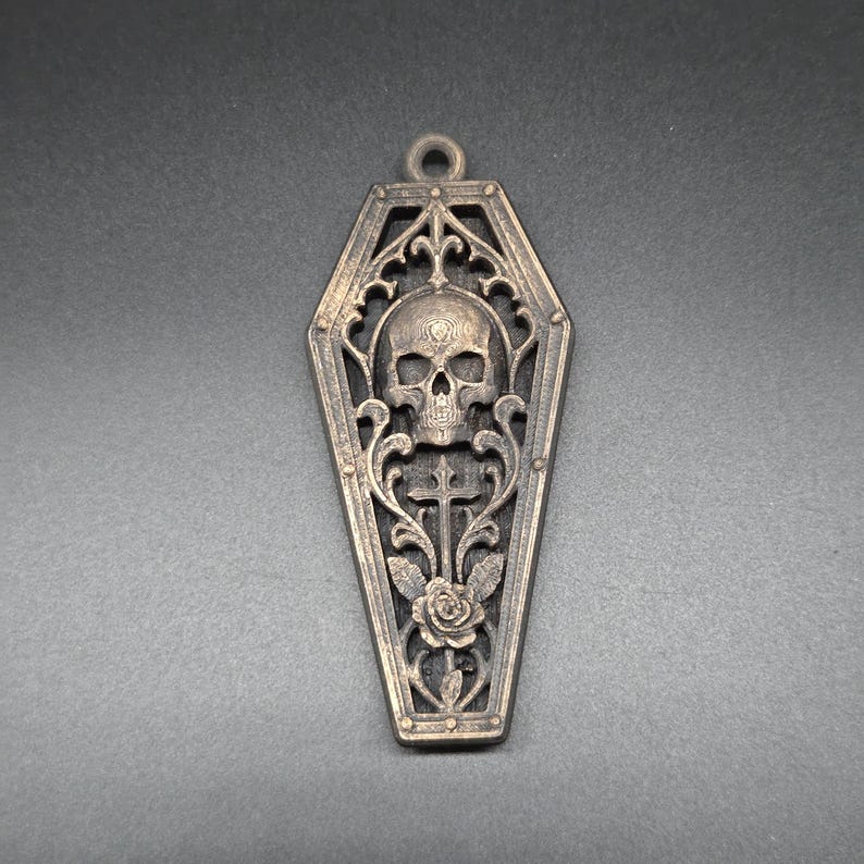 May include: A bronze-toned metal pendant shaped like a coffin, featuring an intricate openwork design. The center displays a detailed human skull positioned above a cross and a blooming rose, all framed by ornate scrollwork and a small hanging loop.