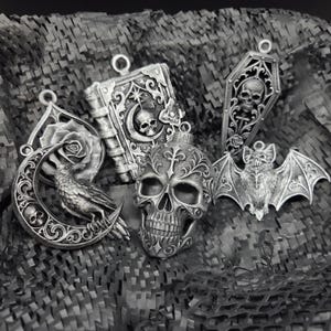 May include: A collection of five silver-toned pendants featuring gothic motifs including a skull, a raven perched on a crescent moon, a book with a skull and moon design, a coffin with a skull, and a bat with spread wings, all with intricate details.