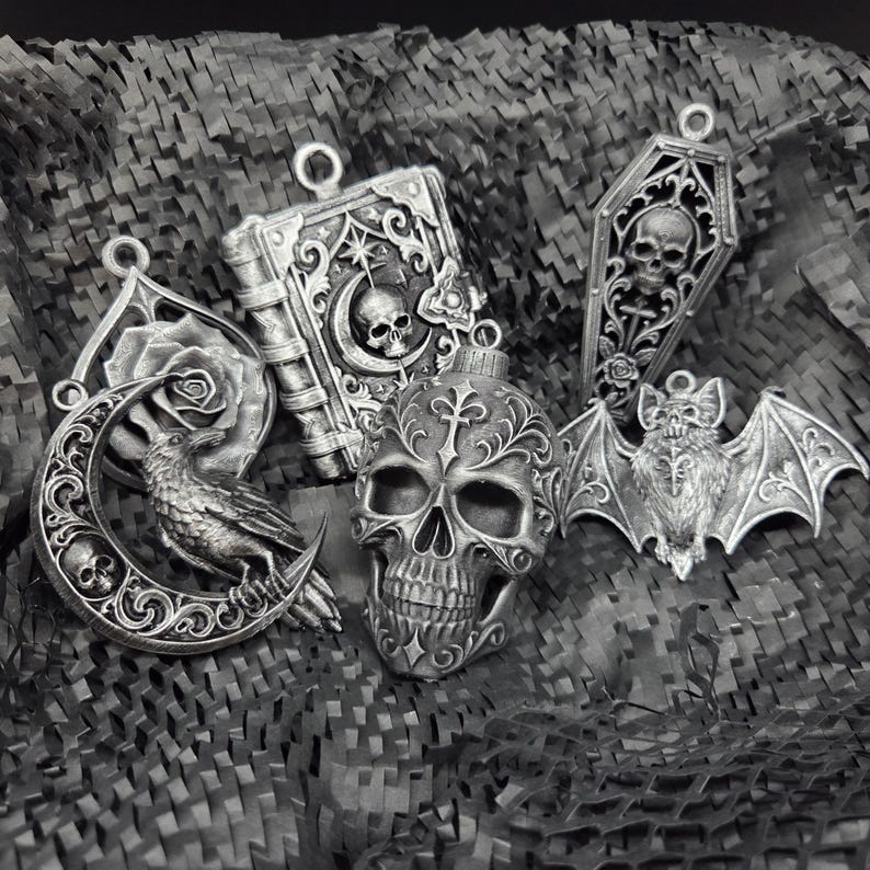 May include: A collection of five silver-toned pendants featuring gothic motifs including a skull, a raven perched on a crescent moon, a book with a skull and moon design, a coffin with a skull, and a bat with spread wings, all with intricate details.