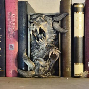Può includere: Un paio di ferma libri in argento a forma di mostro ringhioso con denti affilati e un corno lungo e ricurvo. I ferma libri sono posizionati tra due libri su uno scaffale.