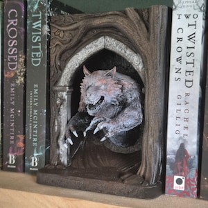 Op de afbeelding: Een boekenlegger met een weerwolf die uit een stenen boog komt. De boekenlegger is gemaakt van geverfd hars en heeft een donkerbruine houtlook. De weerwolf is grijs en wit met rode ogen. De boekenlegger is ontworpen om boeken rechtop te houden.