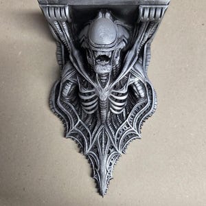 Könnte beinhalten: Eine detaillierte, graue Alien-Themen-Regalhalterung. Die Halterung zeigt ein geformtes Xenomorph-Design mit komplizierten Details, einschließlich des Kopfes der Kreatur, des Brustkorbs und einer dekorativen, spitzen Basis. Das Regal wird vom Körper des Aliens getragen.