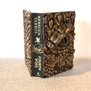 May include: A brown, textured book titled "Fourth Wing" by Rebecca Yarros. The spine is black with gold lettering. The cover features a detailed, carved design, giving a unique appearance.