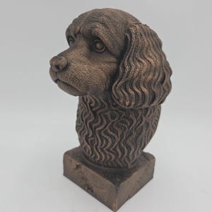 Op de afbeelding: Een bronzen hondenbuste sculptuur. De kop van de hond is naar links gedraaid, met gedetailleerde vacht en gelaatstrekken. De sculptuur staat op een vierkante basis, met een klassiek en elegant ontwerp.