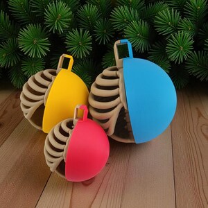 Op de afbeelding: Drie kerstballen met een uniek ontwerp. Elke bal heeft aan de ene helft een skeletribbenkast en aan de andere kant een effen kleur. De kleuren zijn geel, blauw en rood. De ballen liggen op een houten oppervlak met een achtergrond van groene dennennaalden.