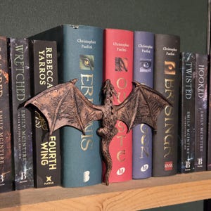 Op de afbeelding: Een bronzen draak boekensteun met uitgespreide vleugels staat voor een rij boeken. De boeken hebben titels als "Eragon", "Brisingr" en "Fourth Wing". De boekensteun voegt een fantasiethema toe aan de boekenplank.