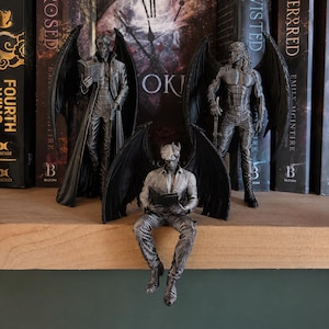 Peut inclure: Trois figurines grisâtres en forme de pierre représentant des humanoïdes ailés. Deux sont debout, l'un lisant un livre et l'autre tenant une épée, tandis que le troisième est assis sur le bord d'une étagère en lisant. Chaque personnage présente des ailes détaillées, des vêtements texturés et des traits du visage définis, avec une finition monochrome.