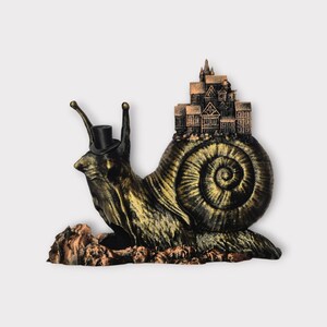 May include: A whimsical sculpture of a snail with a top hat, carrying a miniature town on its back. The snail is bronze and gold, with a detailed shell. The town features small buildings and a castle.