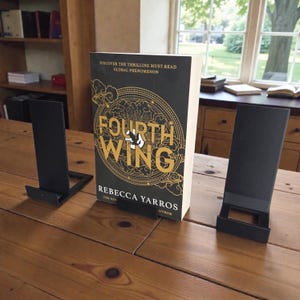 May include: A hardcover book titled "Fourth Wing" by Rebecca Yarros, displayed on a wooden table. The book has a black cover with gold lettering and a circular design. Two black metal book stands flank the book, suggesting a reading setup.