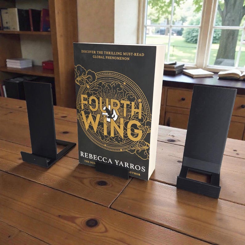 May include: A hardcover book titled "Fourth Wing" by Rebecca Yarros, displayed on a wooden table. The book has a black cover with gold lettering and a circular design. Two black metal book stands flank the book, suggesting a reading setup.