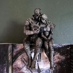 Peut inclure: Une figurine argentée représentant deux personnages assis ensemble, l'un lisant un livre ouvert tandis que l'autre s'appuie contre lui. Les personnages portent des vêtements texturés et détaillés, posés sur une surface ornée d'un cadran d'horloge.