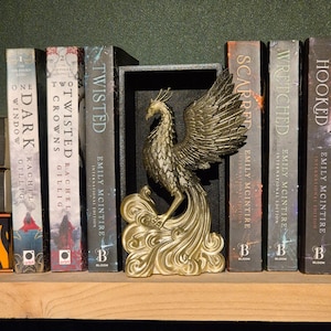 Op de afbeelding: Een gouden phoenix-boekenlegger met een zwarte basis. De boekenlegger houdt een stapel boeken vast met titels als "Twisted", "Scarred" en "Wrecked".