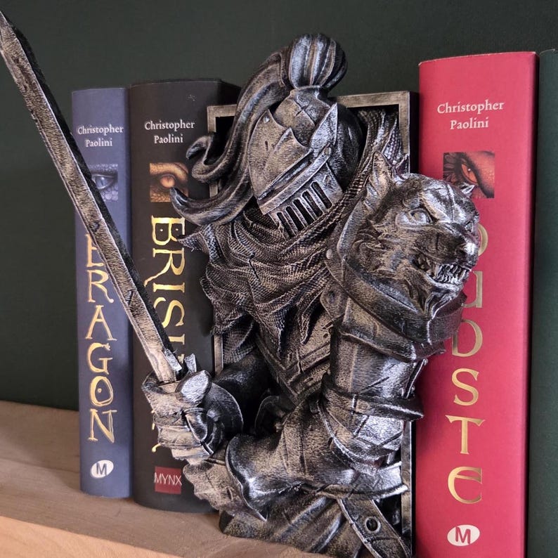 May include: A silver-colored knight bookend with a wolf head design. The knight is holding a sword and is positioned between several books. The books have titles by Christopher Paolini.