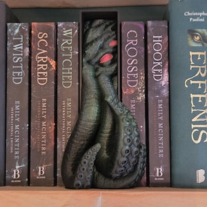 Op de afbeelding: Een donkergroene boekensteun in de vorm van een tentakel met rode accenten, geplaatst tussen boeken. De boeken hebben titels als "Twisted", "Scarred" en "Crossed". De boekensteun voegt een uniek, thematisch element toe.