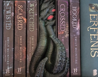 May include: A dark green tentacle-shaped bookend with red accents, placed between several books. The books have titles like "Twisted," "Scarred," and "Crossed." The bookend adds a unique, thematic touch to a bookshelf.