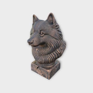 Op de afbeelding: Een bronzen wolfskopsculptuur. De sculptuur heeft een gedetailleerde vachttextuur en staat op een vierkante sokkel. De kop van de wolf is iets opzij gedraaid.
