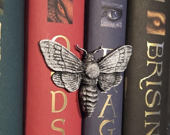 May include: A silver-colored moth-shaped book clip is attached to the spine of a red book. The book is next to other books with dark covers. The clip has detailed wing and body patterns.