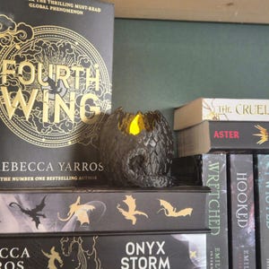 Op de afbeelding: Een zwarte draken-eivormige kandelaar met een geel LED-licht erin, op een plank met boeken. De boektitels zijn onder meer "Fourth Wing" en "Onyx Storm". De kandelaar heeft een getextureerd oppervlak.