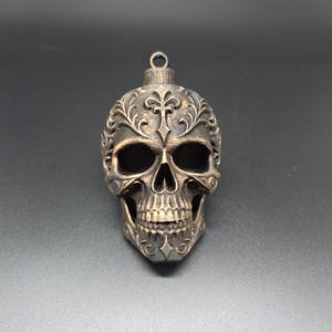May include: A bronze-colored skull ornament featuring intricate embossed floral and cross patterns across the forehead and cheekbones. The skull has deep, hollow eye sockets and a detailed jawline, topped with a small circular metal hanging loop.