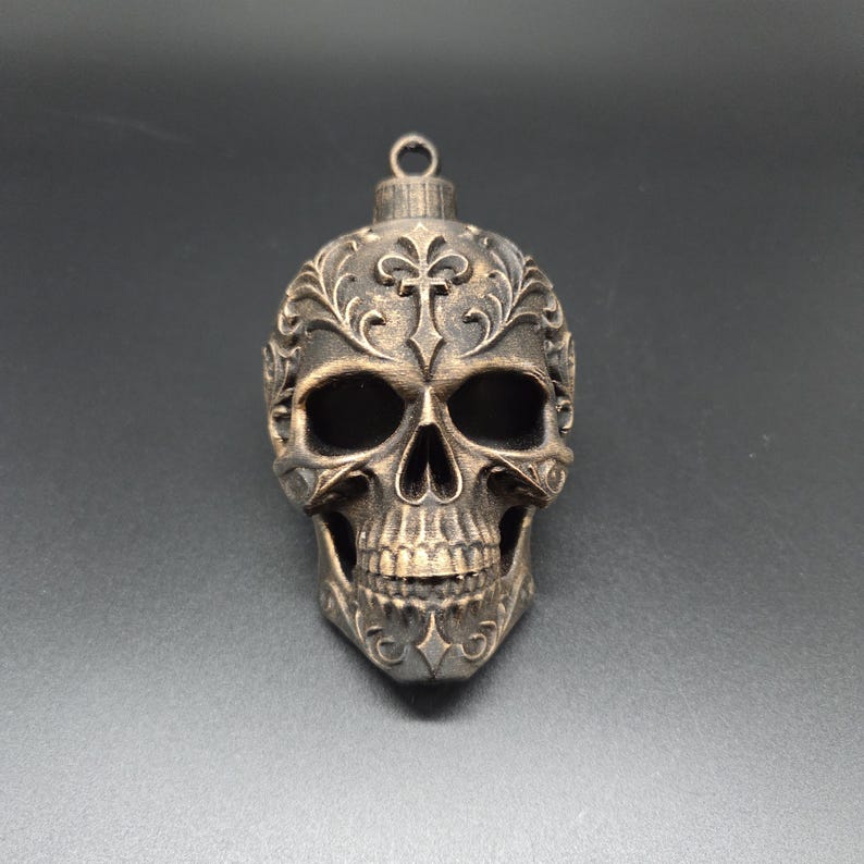 May include: A bronze-colored skull ornament featuring intricate embossed floral and cross patterns across the forehead and cheekbones. The skull has deep, hollow eye sockets and a detailed jawline, topped with a small circular metal hanging loop.