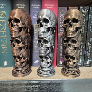 Puede incluir: Tres esculturas decorativas de calaveras apiladas, en tonos bronce, plata y oro. Las esculturas se exhiben en una estantería frente a una fila de libros.