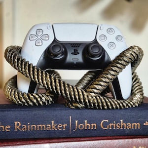 Puede incluir: Un mando de PlayStation 5 blanco está sujeto por un soporte de cuerda dorada. El mando está apoyado en un libro titulado "The Rainmaker" de John Grisham.