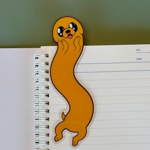May include: A yellow cartoon character bookmark in the shape of a long, wavy dog, with a happy expression. The bookmark is placed on the top of a spiral-bound notebook with lined pages. The background is a solid green color.