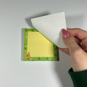 May include: A square sticky note pad with a yellow center and a green border with a triangle pattern. A small illustration of a money bag is in the corner. A white sheet is being peeled back, revealing the yellow sticky notes.
