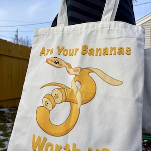 Könnte beinhalten: Weißer Canvas-Tote-Bag mit einem humorvollen Motiv einer Schlange, die sich um eine Bananenschale und Bananenscheiben windet. Der Text "Are Your Bananas Worth it?" ist in Gelb gedruckt. Die Tasche hat weiße Träger.