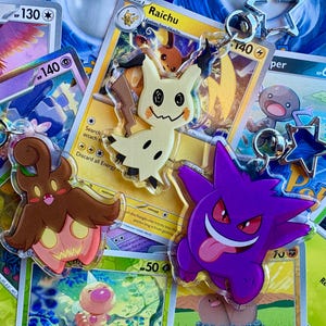 May include: A collection of Pokémon trading cards and acrylic keychains. The keychains feature Mimikyu, Gengar, and a brown and pink character. The cards display various Pokémon, including Raichu and Tinkaton.