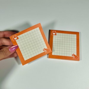 May include: Two orange square sticky notes with a grid pattern and pixelated frog illustrations. One note is held by a hand. The notes have a white grid pattern and frog illustrations in the corners.