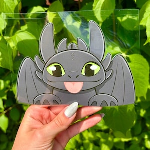 May include: A die-cut vinyl sticker featuring a dark gray dragon character with large green eyes, pointed ears, and a small pink tongue sticking out. The dragon has rounded wings and paws visible at the bottom, presented in a clear protective sleeve.