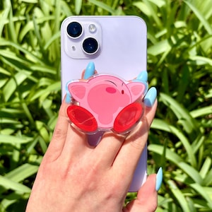 May include: A pink Kirby phone grip on a lilac phone. The grip shows a cartoon Kirby design with red feet and a clear edge. The phone has a dual-lens camera and is held by a hand with blue fingernails.