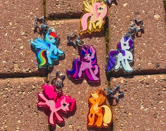 May include: A collection of six acrylic keychains featuring colorful cartoon ponies, each with a silver star-shaped clasp. The characters include a yellow pony with pink hair, a blue pony with a rainbow mane, a purple pony, a white pony, a pink pony, and an orange pony.