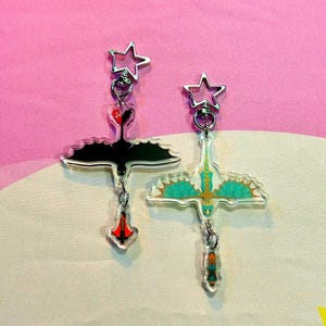 May include: Two acrylic dragon-shaped keychains. One is black with red accents, the other is teal and yellow. Both have star-shaped clasps and dangling charms. The keychains are displayed against a pink and white patterned background.