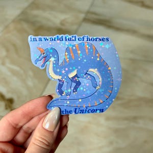 Può includere: Un adesivo olografico raffigurante una creatura blu simile a un dinosauro con strisce arancioni e un corno. L'adesivo presenta il testo "In a world full of horses" e "the Unicorn". Lo sfondo è un gradiente blu con brillantini a forma di stella.