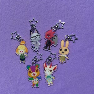 Könnte beinhalten: Eine Sammlung von sieben Schlüsselanhängern aus Acryl mit bunten Zeichentrickfiguren. Jeder Schlüsselanhänger hat einen sternförmigen Verschluss. Die Figuren umfassen einen Hund, eine Katze, ein rotes teufelähnliches Wesen, ein Kaninchen, einen Bären und eine Ziege.