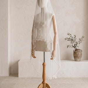 Pode incluir: Um véu de noiva branco transparente sobre um manequim, com um design de duas camadas e delicados adornos brancos em forma de gota espalhados pela camada superior e contornando as bordas festonadas do tecido de tule.