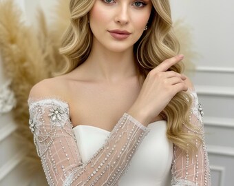 May include: A white off-the-shoulder wedding dress with long, sheer sleeves. The sleeves are adorned with intricate beadwork and crystal embellishments. The model has blonde hair and is wearing makeup.