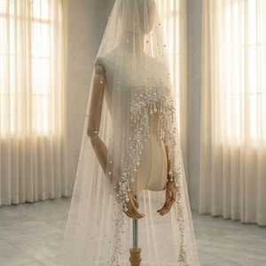 May include: A sheer, ivory-colored bridal veil with delicate pearl embellishments. The veil is draped over a mannequin, showcasing the cascading pearl strands that adorn the edges. The pearls add a touch of elegance and sophistication to the bridal accessory.