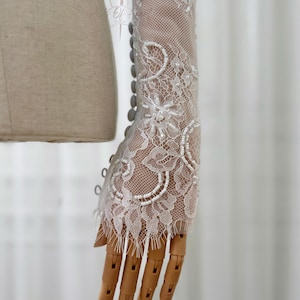 May include: Close-up of a white lace sleeve with floral and beaded details. The sleeve is attached to a mannequin arm with a wooden hand. The lace features intricate patterns and small pearl-like beads. Buttons run along the side of the sleeve.